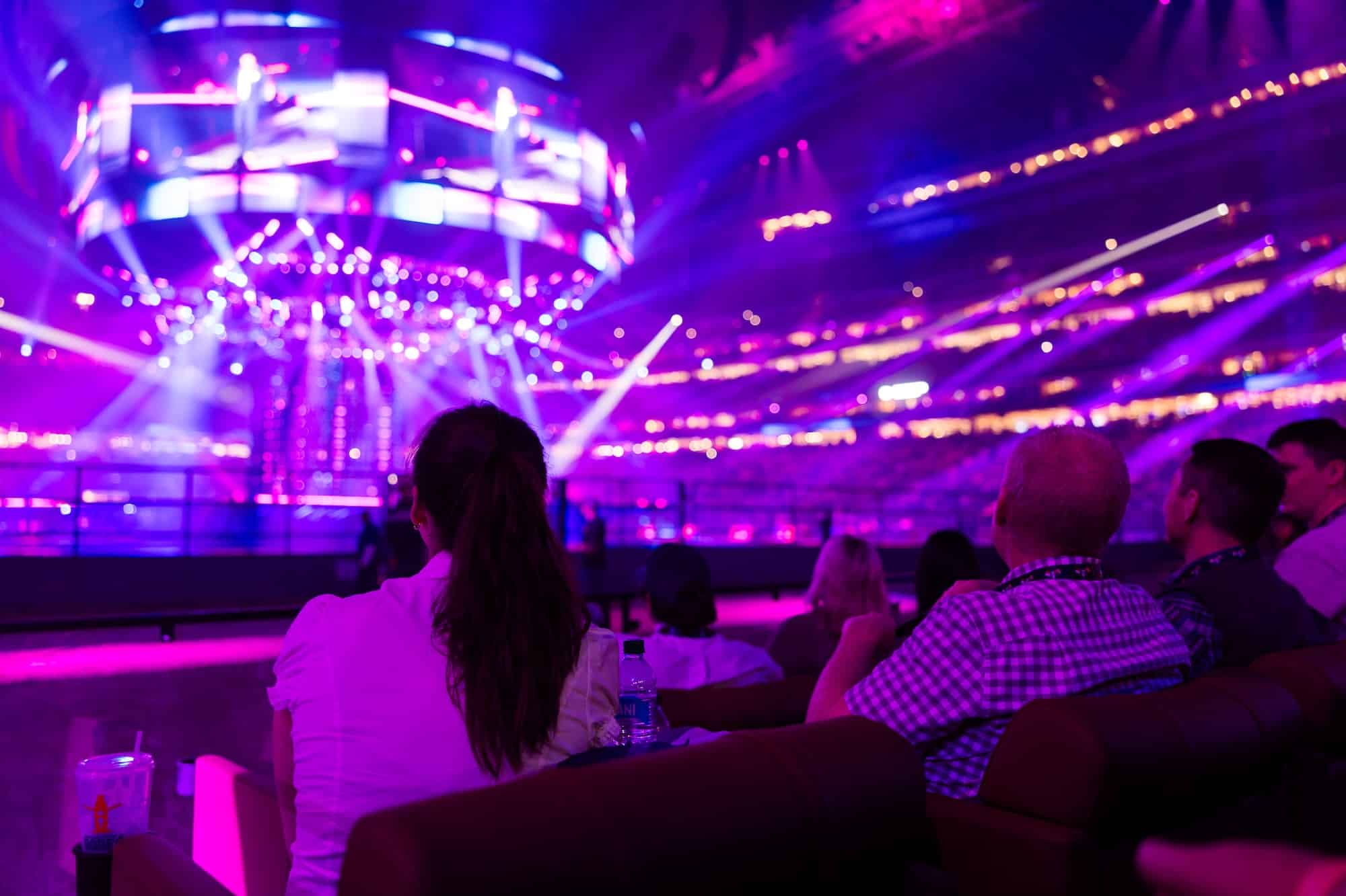 Audience seated in a dark arena bathed in purple lights watching a circular, multi‑screen stage with bright spotlights above the performance area cromatic glow.