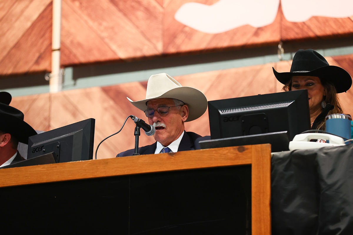 A man in a white cowboy hat speaks into a microphone at an auction, with two computers in front of him and a woman in a black hat nearby.
