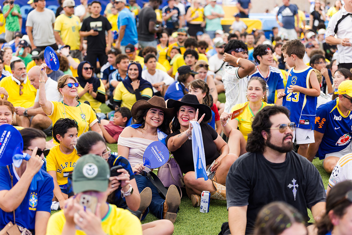 A large crowd of soccer fans, many wearing yellow and blue Brazil jerseys, sit on a grass field at an outdoor event. In the center, two women wearing cowboy hats smile at the camera.