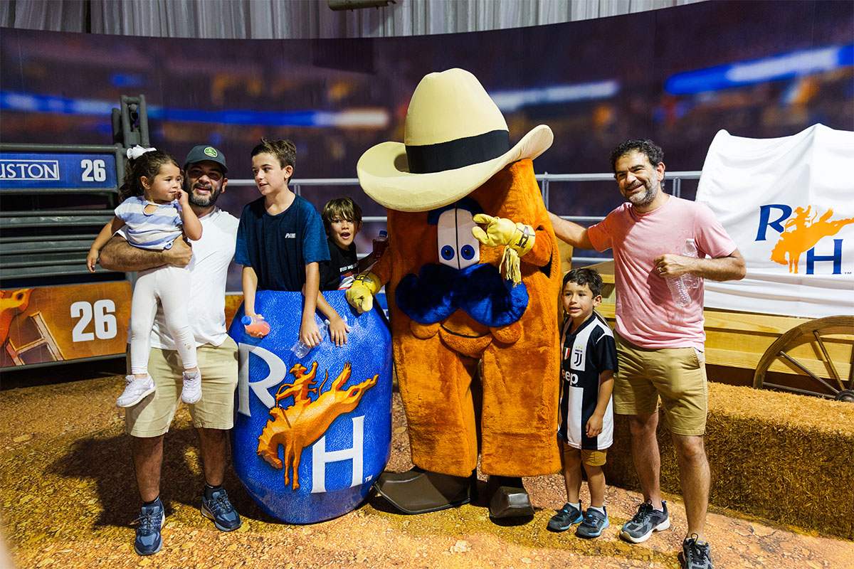 Two men and three children pose for a photo with the RODEOHOUSTON mascot, a large orange character wearing a tan cowboy hat, next to a blue RODEOHOUSTON branded barrel. The group stands on a dirt floor in front of rodeo arena signage and a RODEOHOUSTON logo banner.