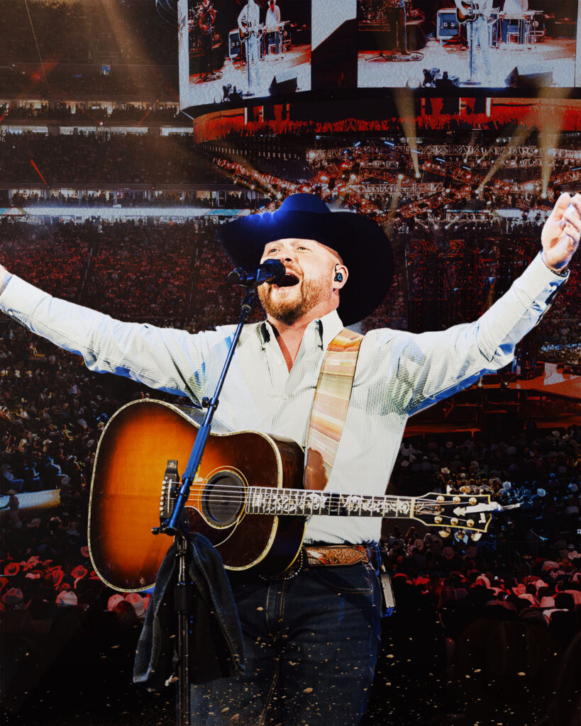 Country musician with a hat and acoustic guitar performing on a large stadium stage, arms raised to the crowd.
