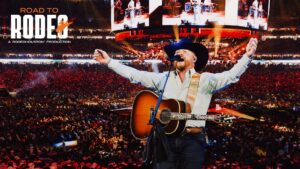 Cowboy singer with guitar on a large arena stage, arms raised, crowd cheering; 'ROAD TO RODEO' banner visible above.