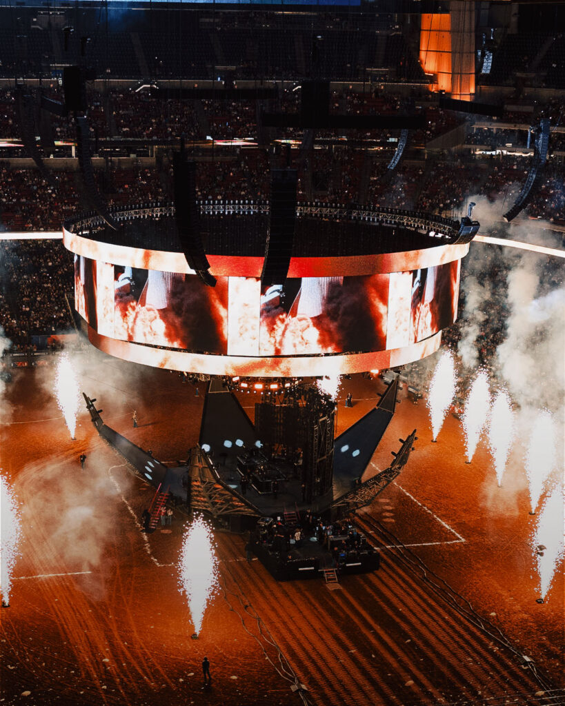 Massive circular stage in a stadium with pyrotechnics firing upward and a cheering crowd surrounding it