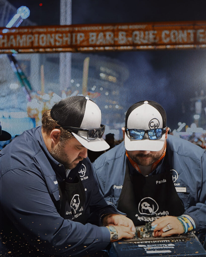 Two men in blue shirts and aprons work on a metal project at a barbecue contest, sparks flying in the foreground.